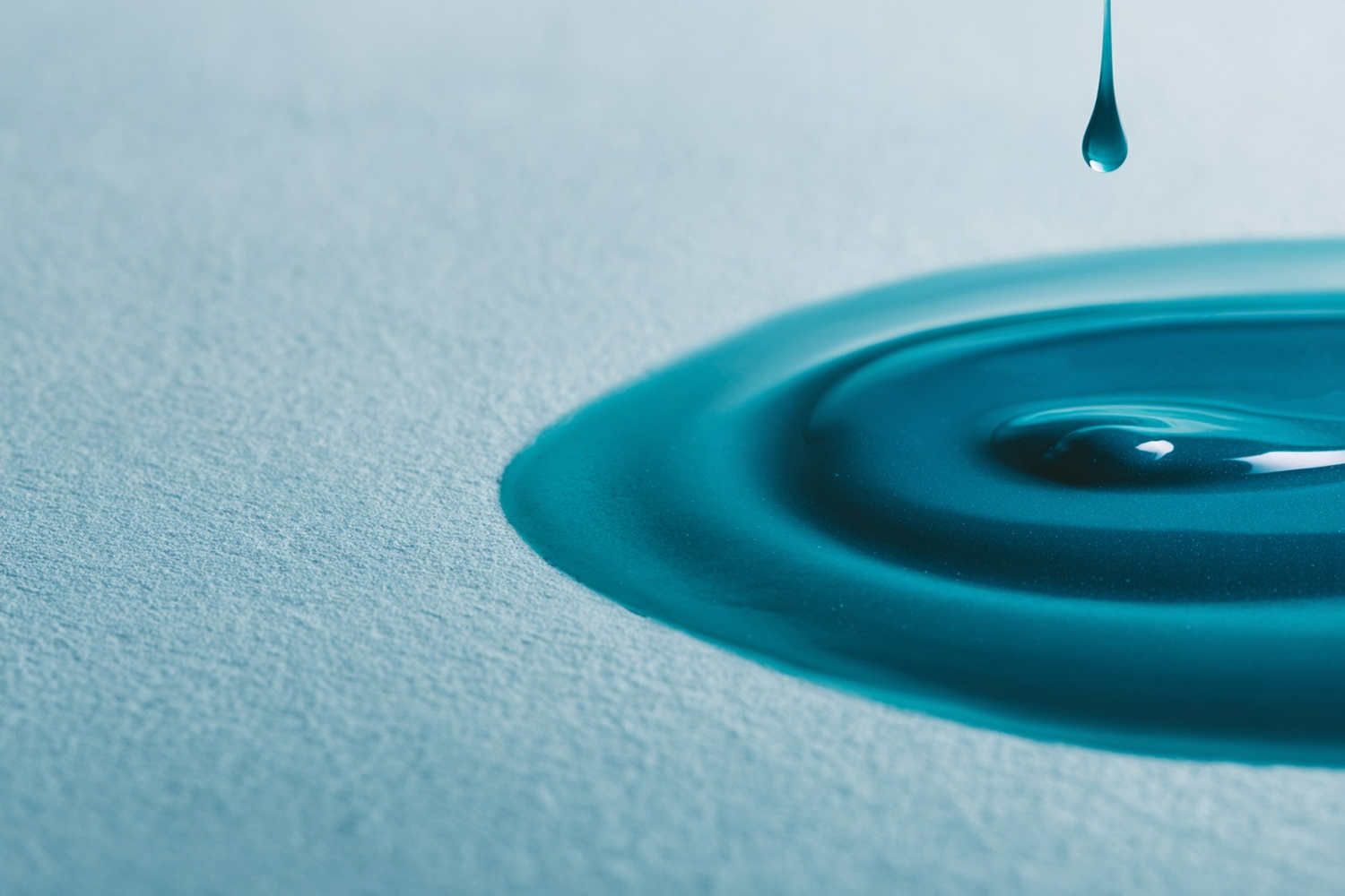 A teal droplet falling into a spreading pool with ripples on a pale surface